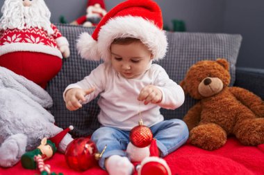 Sevimli beyaz bebek evdeki kanepede oturmuş Noel süsleme balosuyla oynuyor.