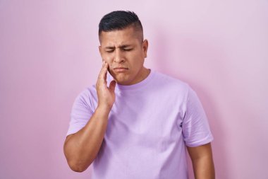 Pembe arka planda duran İspanyol genç adam diş ağrısı ya da diş hastalığından dolayı acı dolu bir ifadeyle ağıza dokunuyor. dişçi 