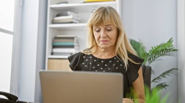 Ciddi ama rahat, güzel orta yaşlı sarışın iş kadını etrafta patronluk taslıyor. Ofis masasındaki dizüstü bilgisayarla odanın arka planına bakıyorum..