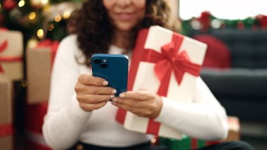 Orta yaşlı İspanyol kadın akıllı telefon kullanıyor. Evdeki Noel ağacının yanında oturuyor.