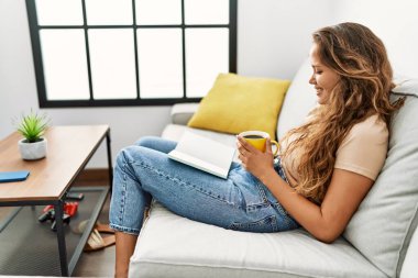 Young beautiful hispanic woman reading book and drinking coffee sitting on sofa at home