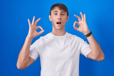 Mavi arka planda dikilen beyaz, sarışın adam şaşırmış ve şaşırmış görünüyor. Parmaklarıyla onay sembolü yapıyor. çılgın ifade 