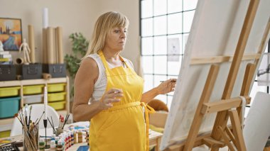 Güzel, orta yaşlı sarışın kadın ressam ciddi ciddi kapalı mekan sanat stüdyosunda resim çizmeye odaklanmış.