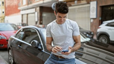 Young hispanic man using smartphone sitting on car at street