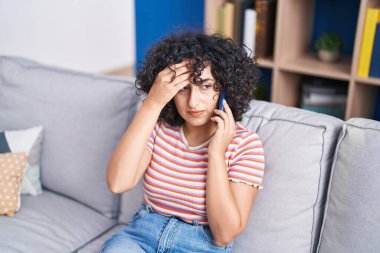 Ortadoğulu genç bir kadın evde endişeli bir ifadeyle akıllı telefondan konuşuyor.