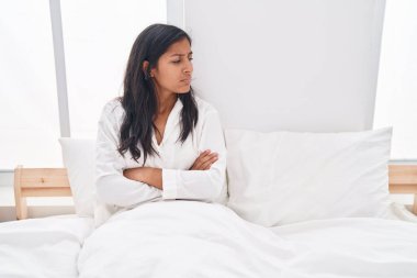 Young beautiful hispanic woman sitting on bed with unhappy expression and arms crossed gesture at bedroom