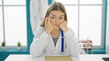 Genç sarışın kadın doktor, klinikte masada yorgun oturuyor.