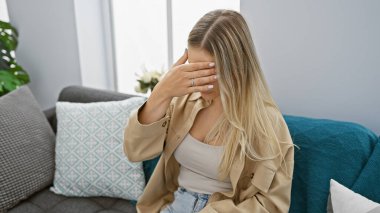 Stresli genç sarışın kadın evdeki kanepede tek başına oturuyor, depresyonla mücadele ederken baş ağrısı ve duygularla boğuşuyor..