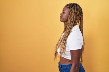 Sarı arka planda dikilmiş örülmüş saçları olan Afro-Amerikalı bir kadın. Yan tarafa bakıyor, rahat poz veriyor ve kendine güvenen bir gülümseme takınıyor.. 