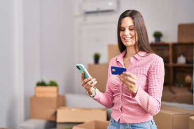 Young beautiful hispanic woman using smartphone and credit card at new home