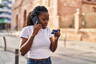 African american woman talking on smartphone holding credit card at street
