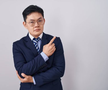 Genç Asyalı takım elbiseli ve kravatlı bir adam el parmağı ile yan tarafı işaret ederek ciddi ve sakin bir yüz ifadesi sergiliyor. 