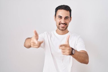 Beyaz arka planda duran yakışıklı İspanyol adam mutlu ve komik yüzlü parmaklarını kameraya doğrultuyor. iyi enerji ve titreşimler. 