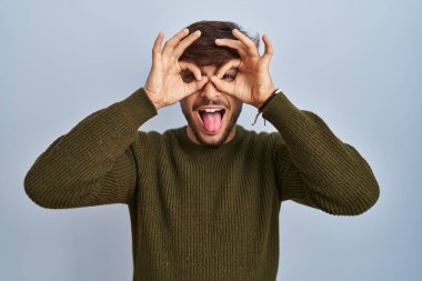 Arab man with beard standing over blue background doing ok gesture like binoculars sticking tongue out, eyes looking through fingers. crazy expression. 