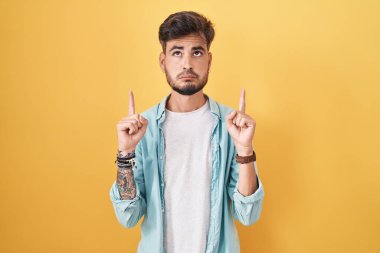 Sarı arka planda dikilen İspanyol dövmeli genç adam üzgün ve üzgün görünüyor, parmaklarıyla yönü gösteriyor, mutsuz ve depresif.. 
