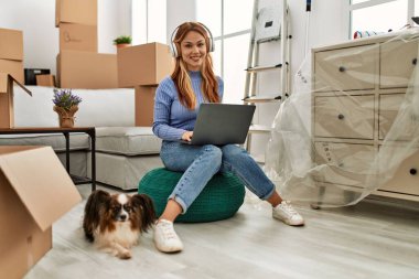 Laptop ve kulaklık kullanan beyaz bir kadın yeni evinde köpekle oturuyor.