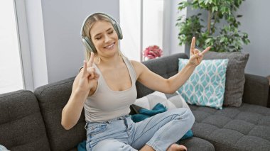 Neşeli genç sarışın kadın kanepede dinleniyor, müzik dinliyor ve evinde tasasız bir şekilde dans ediyor.