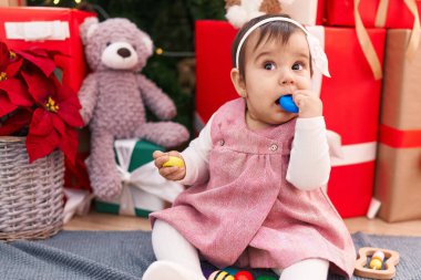 Tapılası İspanyol bebek evde Noel ağacının yanında oturmuş oyuncak yutuyor.