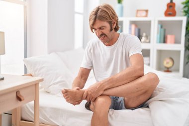 Young blond man suffering for feet injury sitting on bed at bedroom