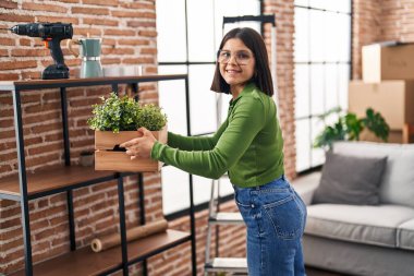 Genç, güzel, İspanyol bir kadın gülümsüyor. Yeni evinde tahta saksı tutuyor.