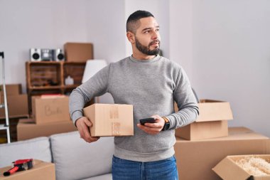 Young latin man using smartphone holding package at new home