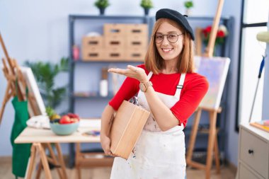 Sanat stüdyosundaki genç kızıl saçlı kadın elinde sanat davasıyla avuç içi açık bir şekilde fotokopi alanını gösteriyor, gülümseyerek mutlu bir şekilde gülümsüyor. 