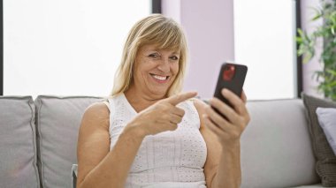 Teknoloji zamanından zevk alan neşeli orta yaşlı sarışın kadın, akıllı telefonu işaret ederken gülümsüyor, evdeki oturma odasındaki kanepede rahatça oturuyor.