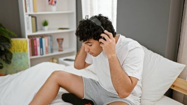 Kulaklık takan genç latin adam yatak odasında dinlenerek müzik dinliyor.
