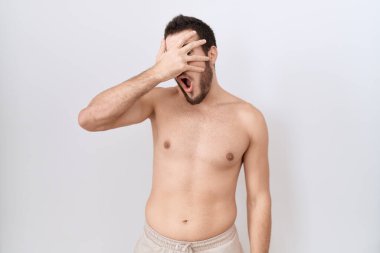 Beyaz arka plan üzerinde üstsüz duran İspanyol genç adam şok içinde yüzünü ve gözlerini kapayarak utanmış ifadesiyle parmaklarının arasından bakıyor.. 