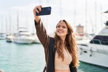 Genç, güzel, İspanyol bir kadın, gülümsüyor. Deniz kenarındaki akıllı telefonun yanında özçekim yapıyor.