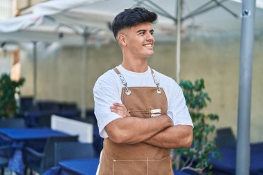 Genç İspanyol garson, gülümsüyor. Kollarını kavuşturup kahve dükkanının terasında duruyor.