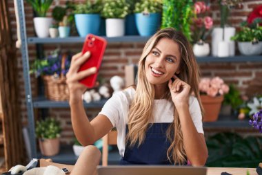 Genç sarışın kadın çiçekçi çiçekçide akıllı telefondan selfie çekiyor.