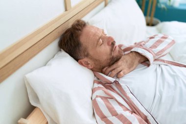 Orta yaşlı bir adam yatakta yatarken boğaz ağrısı çekiyor.
