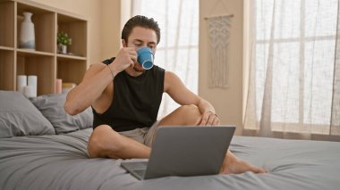 Young hispanic man using laptop drinking coffee at bedroom