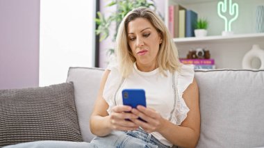 Kanepede oturan akıllı telefon kullanan genç sarışın kadın ciddi bir yüzle evde oturuyor.