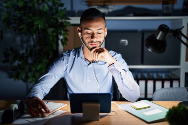 Geceleri ofiste çalışan Latin asıllı genç adam kameraya kollarını kavuşturup gülümseyen ve çenesini kaldıran kendinden emin bir şekilde bakıyor. Olumlu düşünmek. 