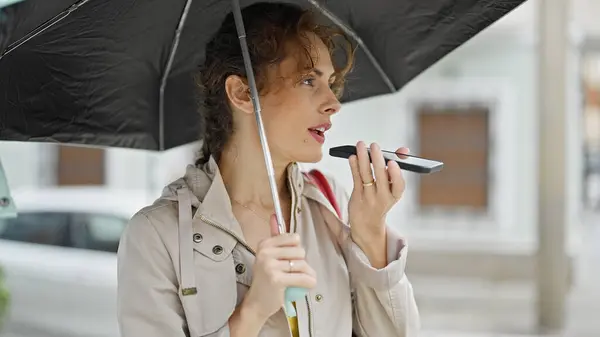 Young woman sending voice message with smartphone holding umbrella at street