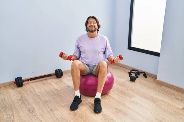 Middle age man training with dumbbells sitting on fit ball at sport center