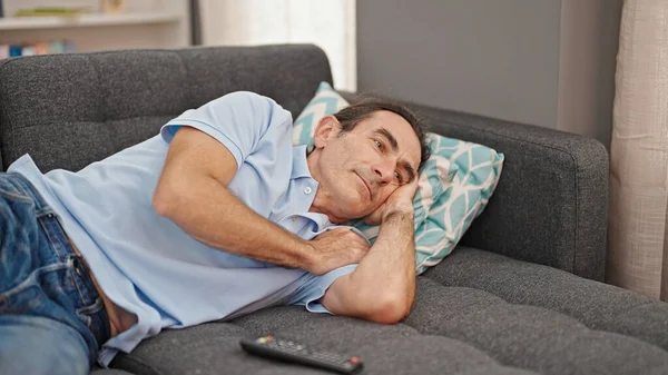 Middle age man watching tv lying on sofa with boring expression at home