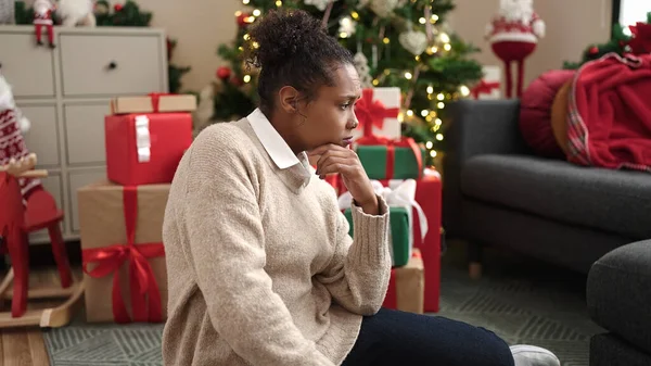 African american woman sitting on floor by christmas tree with sad expression at home