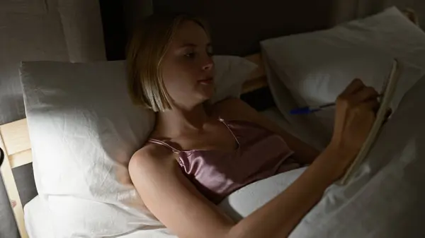 Young blonde woman writing on notebook lying on bed at bedroom