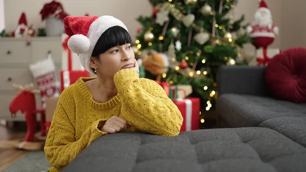 Young chinese woman leaning on sofa by christmas tree with boring expression at home
