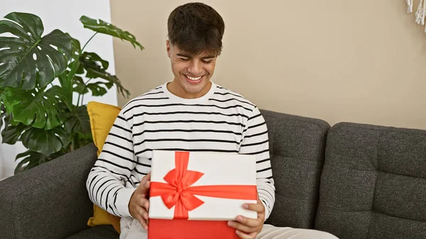 Attractive young hispanic man, sitting comfortably on the sofa at home, smiling with a positive expression as he surprises at unpacking his birthday gift.