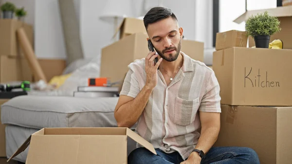 Young hispanic man packing cardboard box talking on smartphone at new home