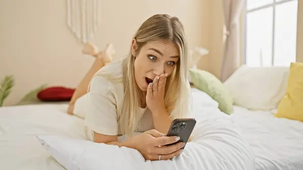 Έκπληκτη Όμορφη Νεαρή Ξανθιά Γυναίκα Ξαπλωμένη Στο Κρεβάτι Χρησιμοποιώντας Smartphone — Φωτογραφία Αρχείου