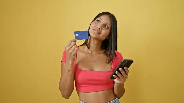年轻的拉丁女人带着智能手机和信用卡购物 对孤立的黄色背景进行思考 — 图库照片