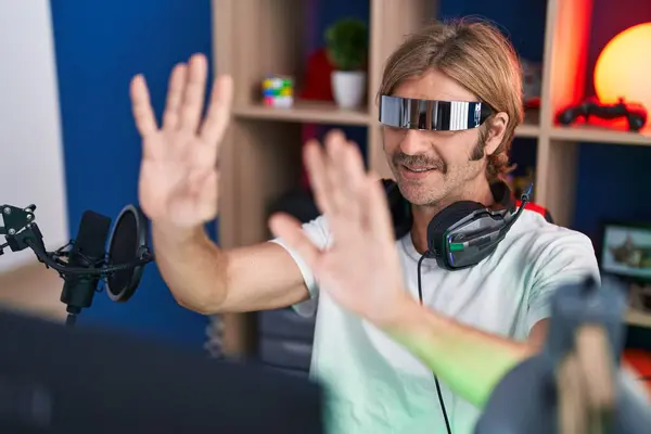 Young blond man streamer playing video game using virtual reality glasses at gaming room