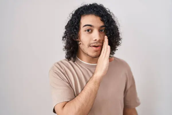 Hispanic man with curly hair standing over white background hand on mouth telling secret rumor, whispering malicious talk conversation