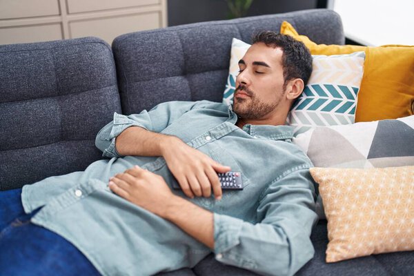 Young hispanic man holding tv remote control lying on sofa sleeping at home