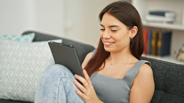 Young beautiful hispanic woman using touchpad sitting on sofa at home